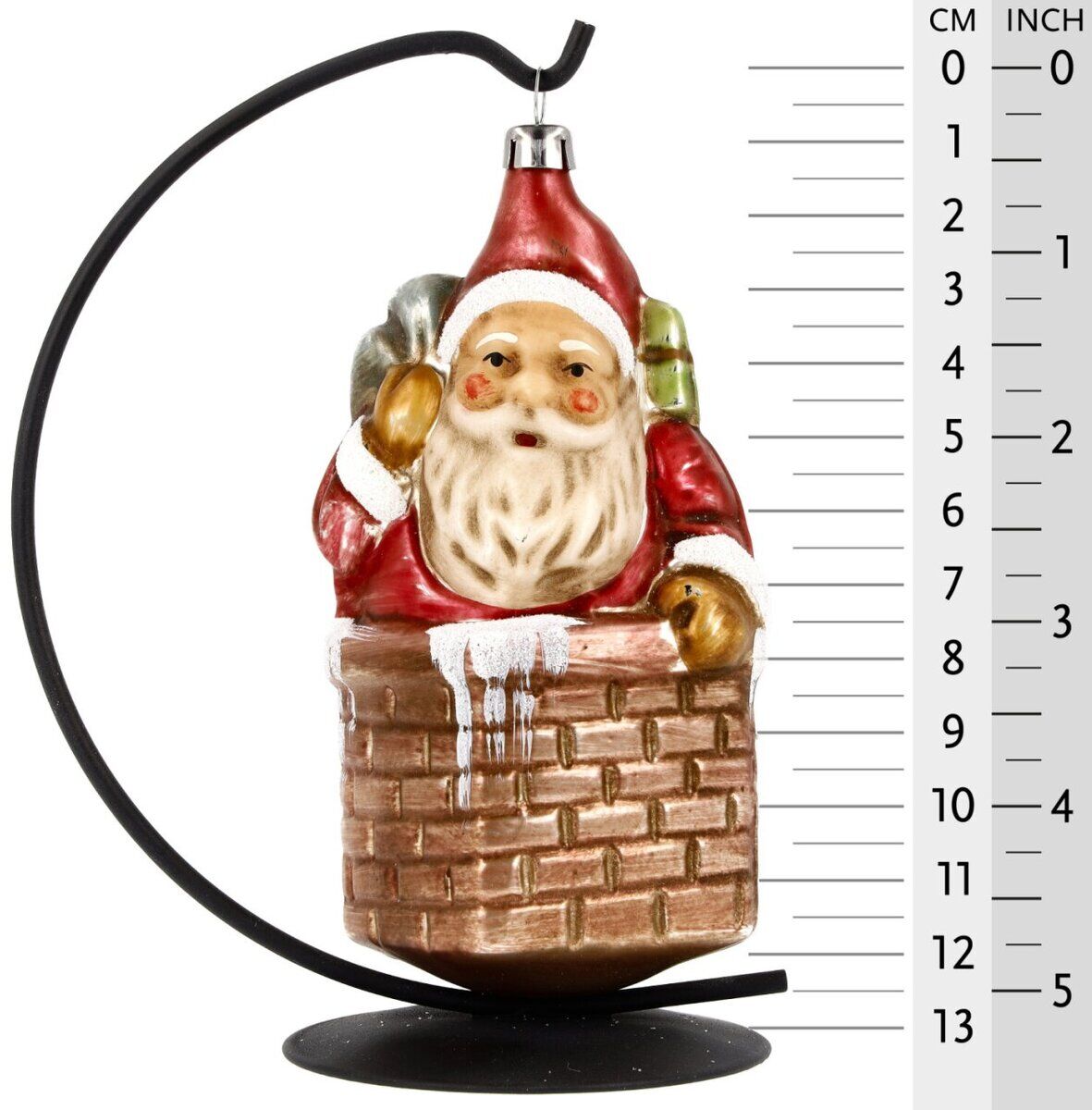 Glasornament in Form eines Nikolaus mit rotem Mantel und Mütze im Schornstein. Der Nikolaus trägt einen Sack auf dem Rücken. Das Ornament ist handbemalt und glänzt. Es hängt an einem metallischen Ständer.