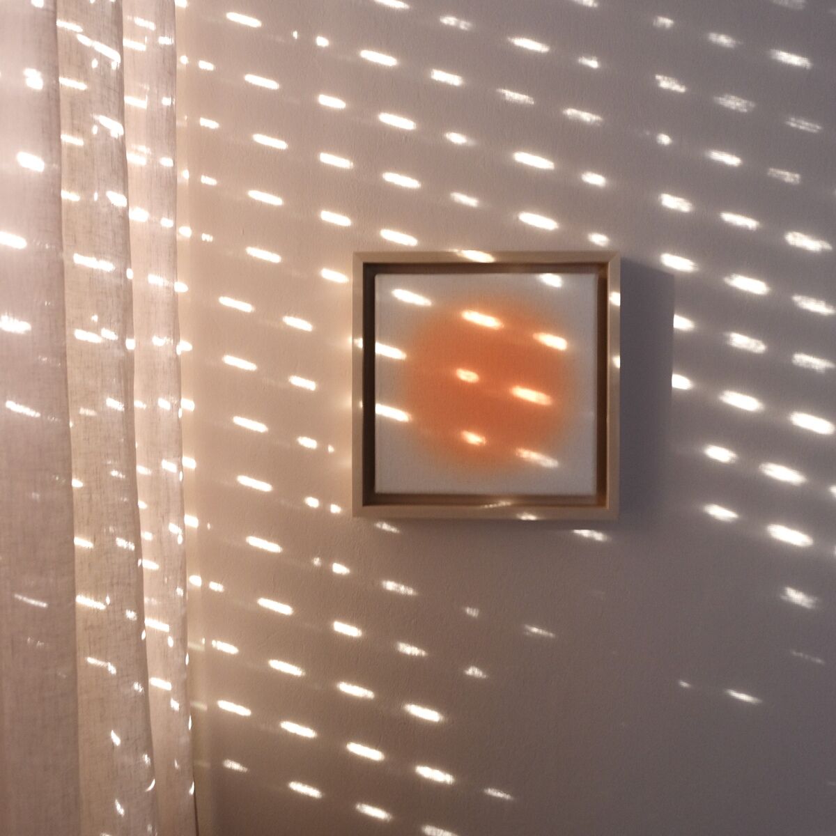 Quadratisches Kunstwerk mit einem orangefarbenen Kreis vor einem weißen Hintergrund, aufgehängt an einer weißen Wand. Sonnenstrahlen erzeugen ein Muster aus Lichtpunkten auf Wand und Kunstwerk. Die Atmosphäre ist ruhig und modern.