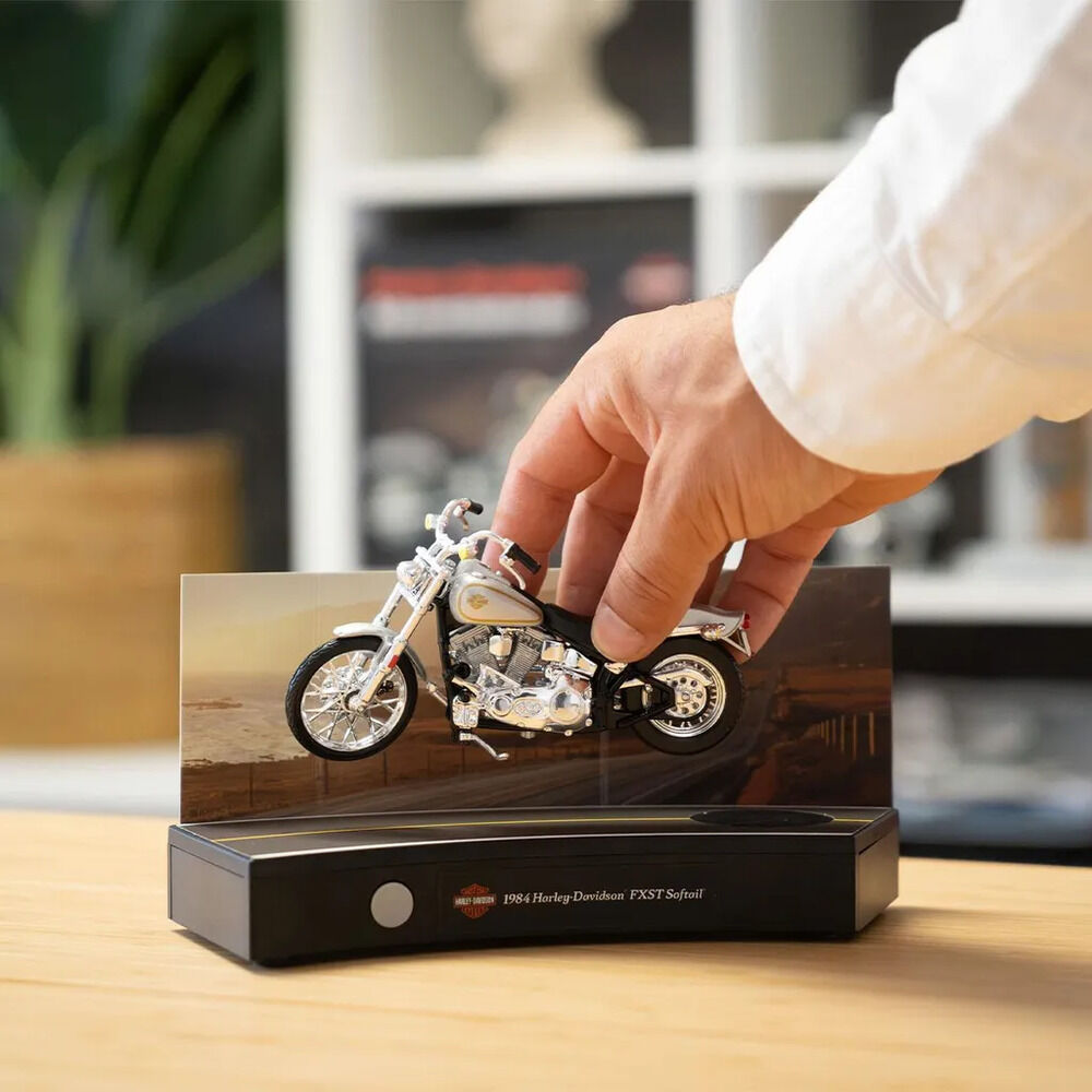 Nahaufnahme eines detailreichen Harley-Davidson-FXST-Softail-Motorradmodells in Schwarz, Silber und Creme auf einer schwarzen Soundbasis. Eine Hand im weißen Hemd setzt das Modell vor eine bedruckte Kulisse; im unscharfen Hintergrund stehen Zimmerpflanze und Regal.