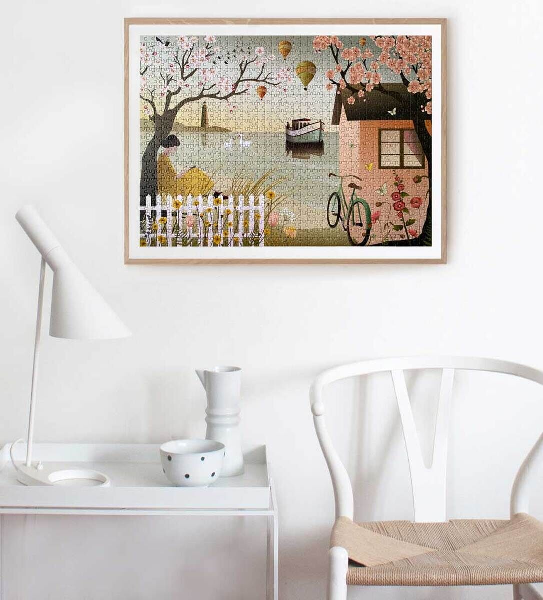 Ein gerahmtes Puzzle an der Wand zeigt eine Frühlingslandschaft am Meer mit einem Baum, einem Haus, einem Fahrrad und Heißluftballons im Hintergrund. Es steht auf einem Tisch neben einem weißen Stuhl.