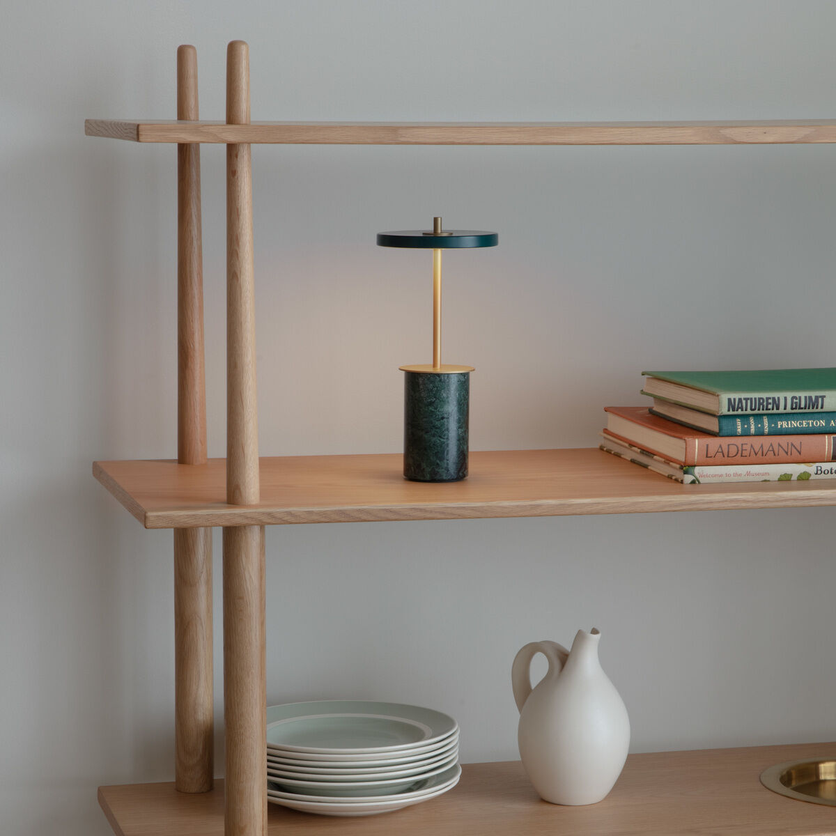 Eine Tischlampe namens Asteria Move Mini steht auf einem offenen Regal aus hellem Holz. Die Lampe hat ein rundes, flaches Leuchtteil, einen schmalen zylindrischen Fuß und dezente goldene Designelemente. Ringsum befinden sich gestapelte Teller, eine weiße Karaffe und Bücher, die das skandinavisch und minimalistisch eingerichtete Wohnzimmer ergänzen.