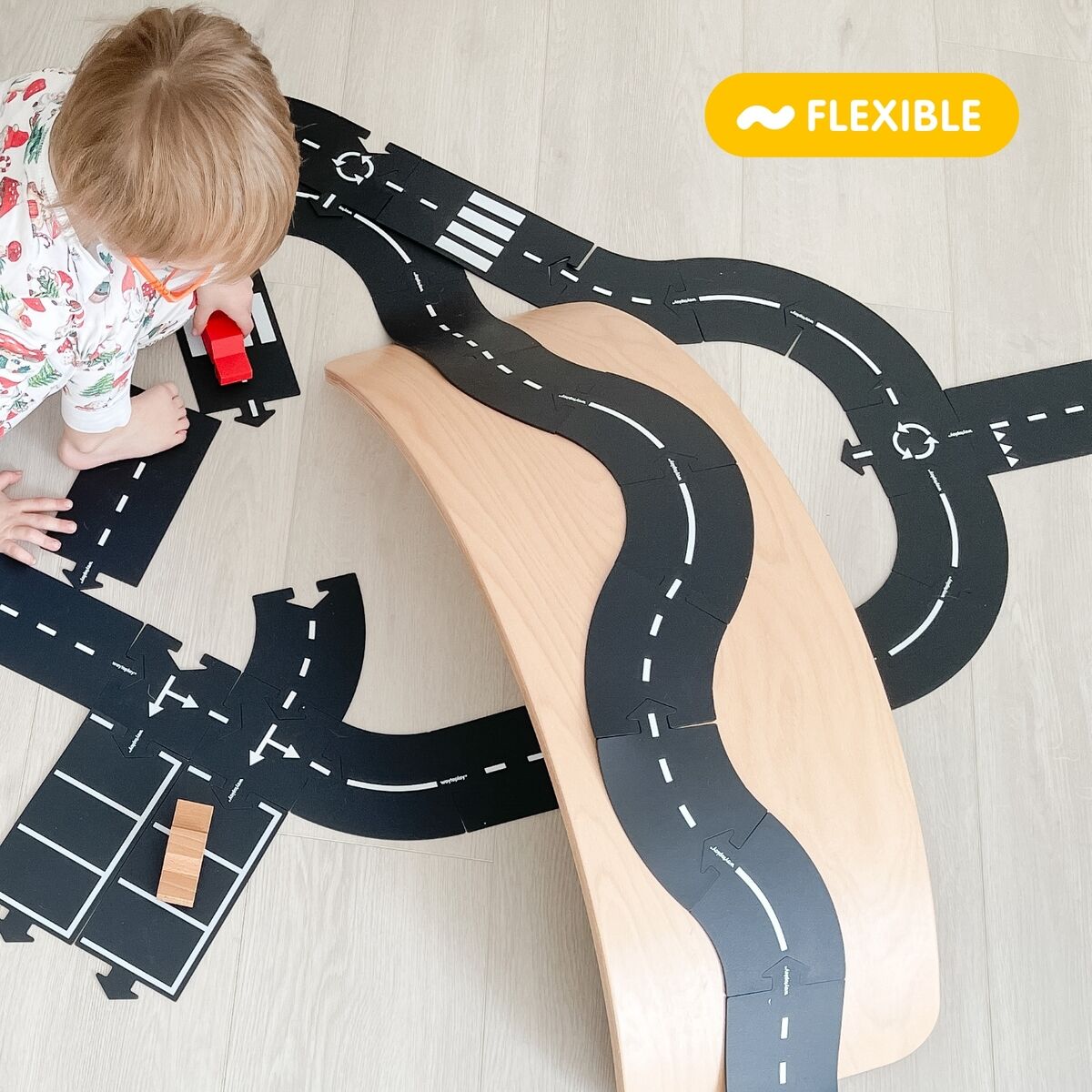 Kleines Kind spielt im Kinderzimmer auf hellem Boden mit einem flexiblen Straßenbau-Set »King of the road«. Mehrere schwarze, modulare Straßenplatten sind ausgelegt, ein Holzauto und eine Holzbrücke ergänzen die Szene. Im Bild befindet sich ein gelbes Schild mit der Aufschrift "flexible".