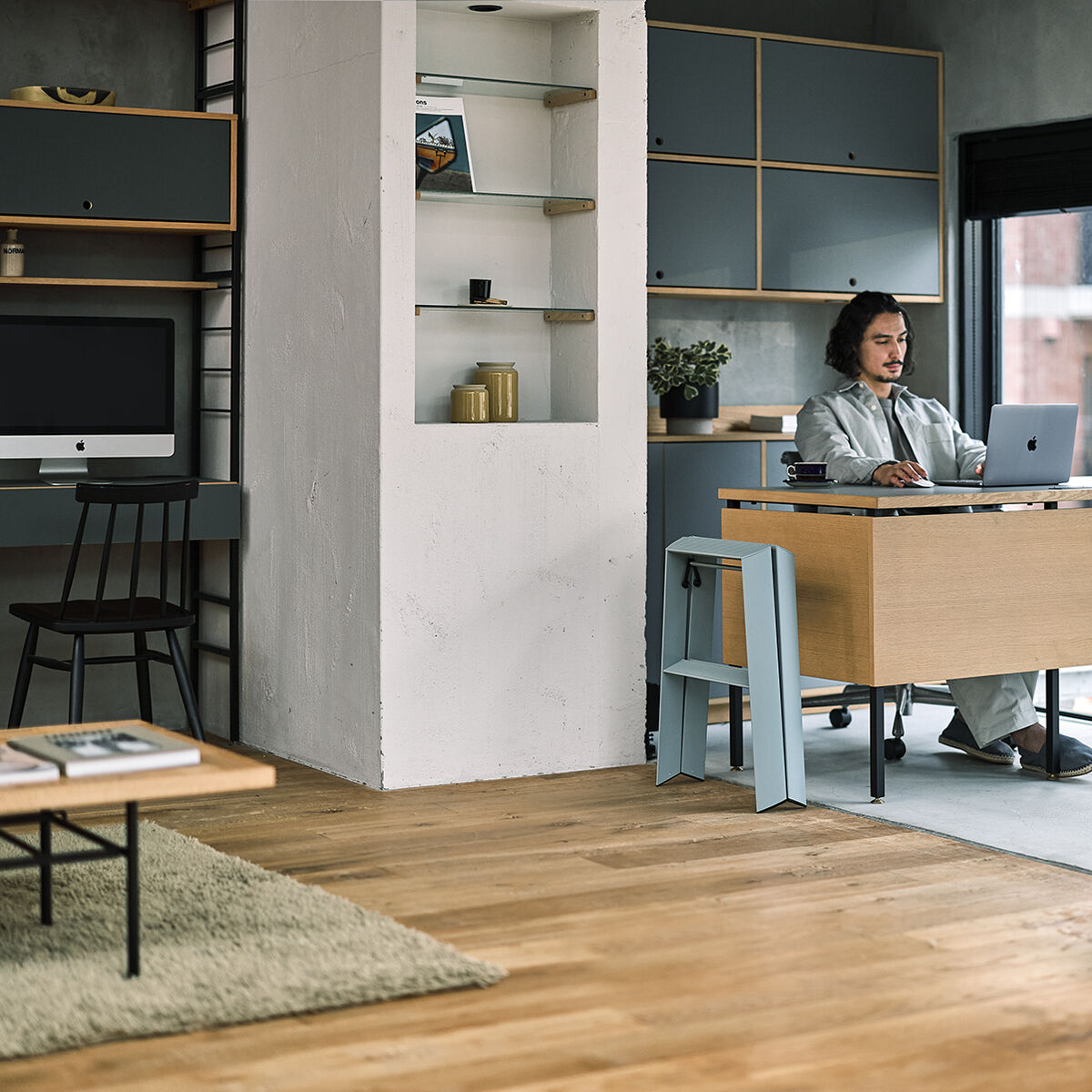 Eine moderne hellblaue Lucano Trittleiter mit zwei Stufen steht in einem stilvoll eingerichteten Büro mit Holzfußboden, hellen Möbeln und zeitgenössischer Einrichtung. Die Trittleiter ist platzsparend neben einem Schreibtisch positioniert und dient als praktisches Wohnaccessoire, um hohe Regale zu erreichen.