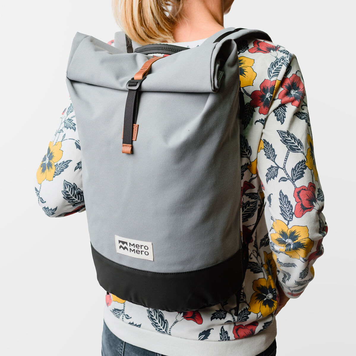 Eine Person trägt einen grauen Rucksack namens 'Wanaka' mit einem schwarzen unteren Teil. Der Rucksack hat einen modernen und schlichten Stil und wird hier von jemandem in einem bunten Sweatshirt präsentiert.