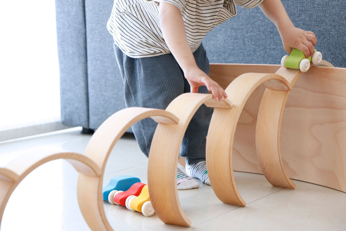 Ein Kind spielt mit einem Regenbogen Holzspielzeug. Das Spielzeug besteht aus natürlich gehaltenen, halbkreisförmigen Holzelementen, die übereinander gestapelt werden. Im Vordergrund liegen zusätzlich bunte, kleine Holzteile. Der Boden ist hell gefliest und im Hintergrund ist ein graues Sofa zu sehen.