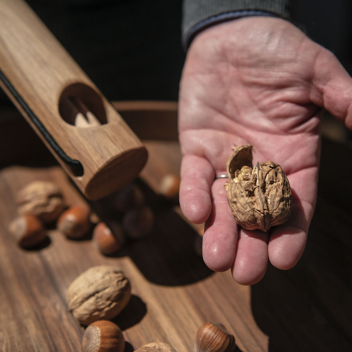 Holz-Nussknacker im Einsatz: Eine Hand hält eine geknackte Walnuss über einer Holzschale mit weiteren Nüssen, während der Nussknacker daneben liegt.