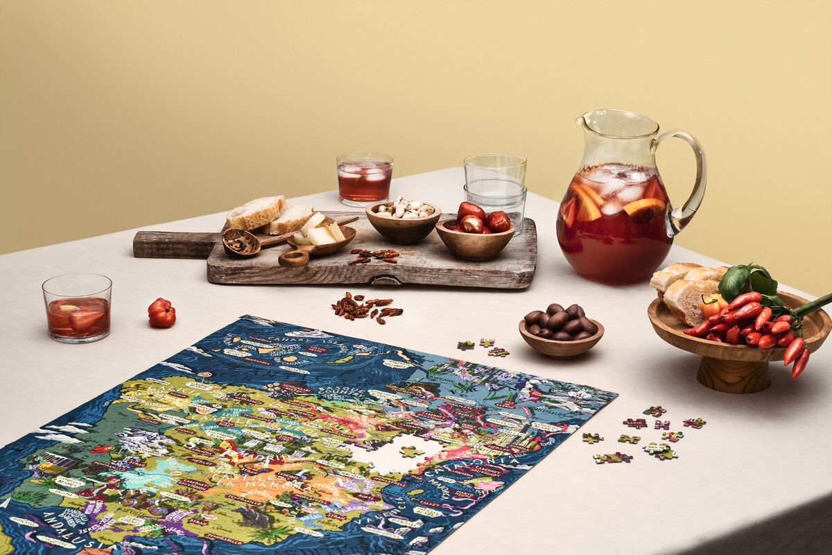 Auf einem Tisch liegt ein Wein-Puzzle mit Spanien-Motiv, umgeben von verschiedenen Snacks wie Brot, Nüssen, Schokolade und einem Krug Sangria. Es gibt Gläser mit Getränken und kleine Schälchen mit weiteren Leckereien. Ein Schneidebrett mit Tomaten, Paprika und Brotstücken ist ebenfalls zu sehen, alles vor einem neutralen Hintergrund.