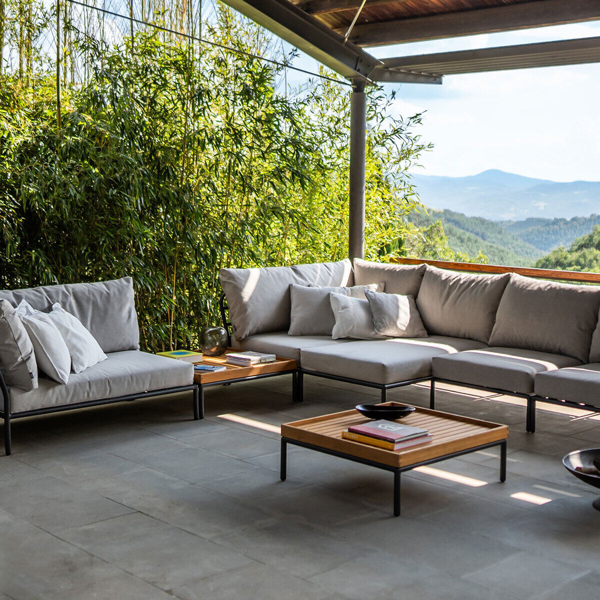 Ein moderner Loungesessel mit Ecksofa, in einem Außenbereich auf einer Terrasse platziert. Die Möbelstücke sind in neutralen Farben wie Grau und Braun gehalten. Im Hintergrund ist eine üppige grüne Landschaft und der blaue Himmel zu sehen, was dem Raum eine entspannte und einladende Atmosphäre verleiht.