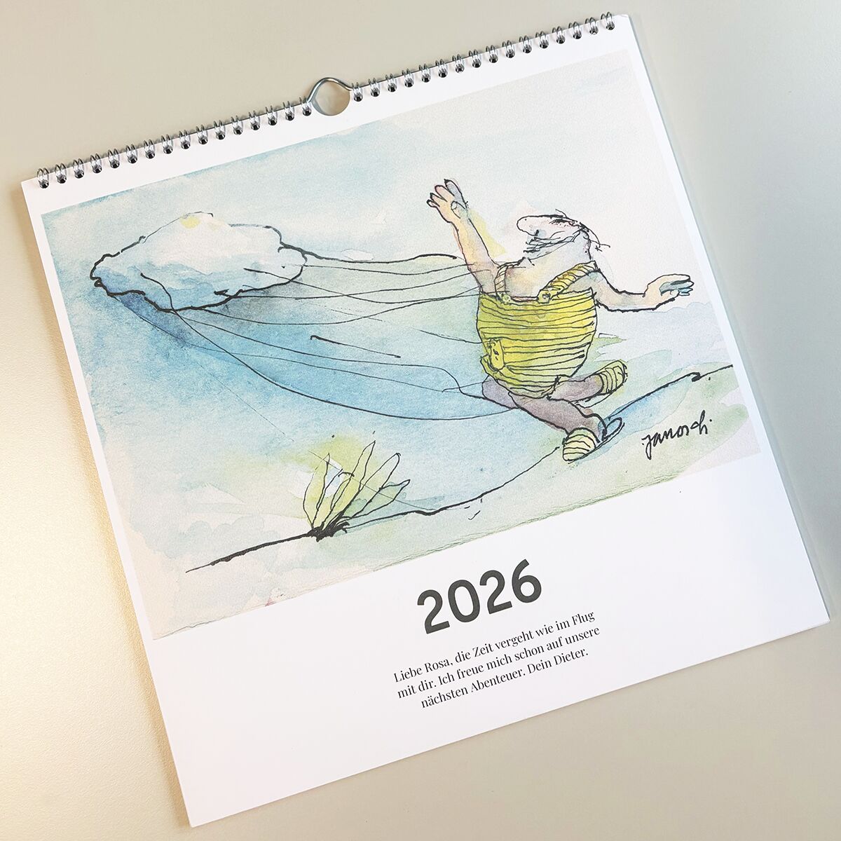 Quadratischer Wandkalender für 2026 mit einer humorvollen, handgezeichneten Illustration im Stil von Janosch. Die Zeichnung zeigt eine fliegende Figur mit flatterndem Umhang in hellen Pastellfarben. Unten befindet sich der Schriftzug '2026' und ein inspirierendes Zitat auf Deutsch. Das Deckblatt liegt auf einer hellen, glatten Oberfläche.