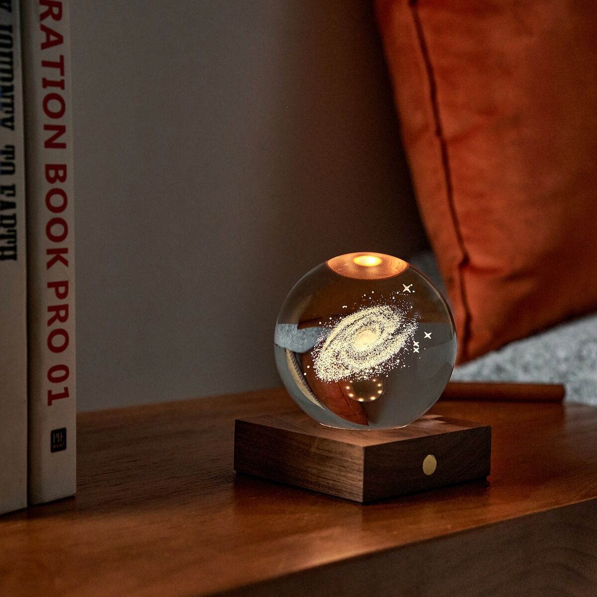 Eine dekorative Tischlampe mit amberfarbenem Licht und einer Kristallkugel, die eine Galaxie darstellt, steht auf einem hölzernen Sockel auf einem Holztisch. Im Hintergrund sind mehrere Kissen und Bücher sichtbar, die eine gemütliche Wohnzimmeratmosphäre schaffen.