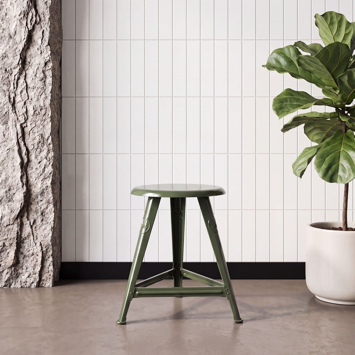 In einem modernen, hellen Raum mit Steinwand und großen, weißen Fliesen steht ein grüner Stahl-Schemel („Just Steel“, Mountain Green von Rowac) mittig am Boden. Rechts daneben steht in einem weißen Topf eine große grüne Zimmerpflanze. Klare Linien und industrielles Design dominieren.