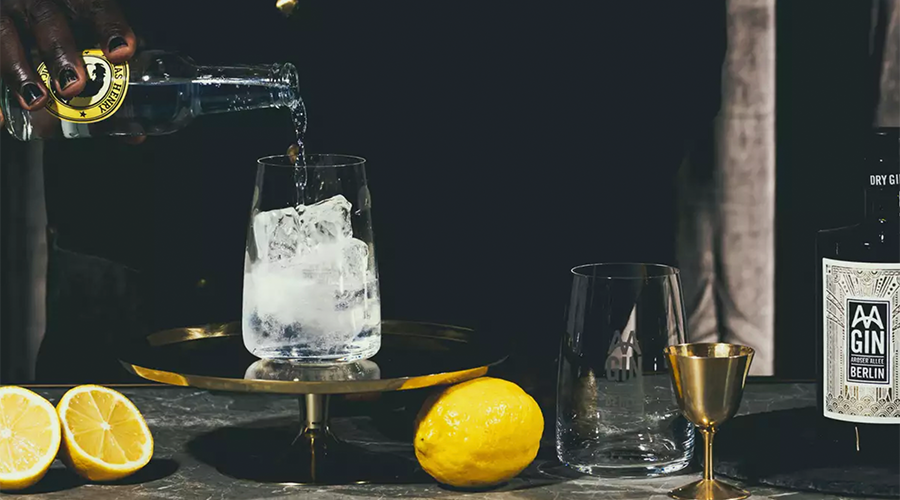Eine Hand gießt Tonic Water in ein Glas mit Eis, umgeben von einer Zitrone, einer Gin-Flasche und einem goldenen Messbecher auf einer dunklen Oberfläche.