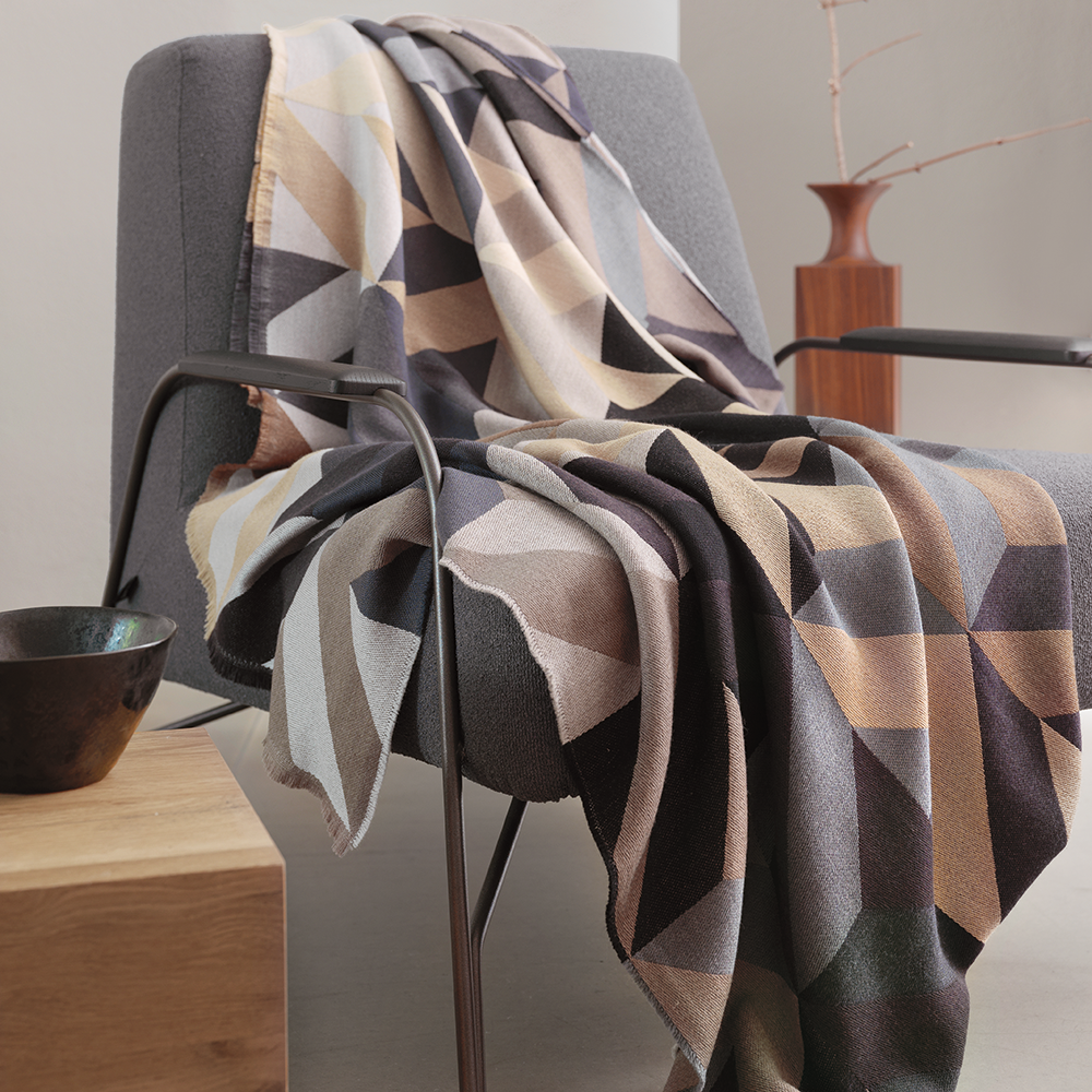 Gemusterte Decke in Erdtönen auf einem modernen Stuhl, daneben eine Holzskulptur und eine Schale.