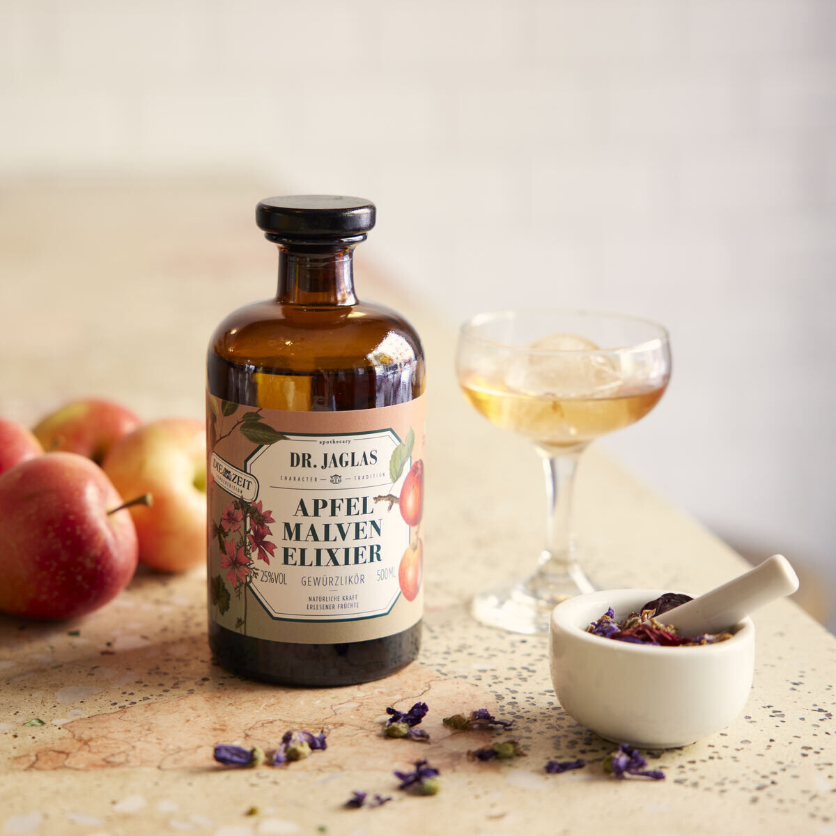 Flasche Apfel Malven-Elixier Dessertlikör von Dr. Jaglas, umgeben von Äpfeln und einem Glas mit Getränk, auf einer rustikalen Oberfläche. Daneben ein kleiner Behälter mit einem Mörser, gefüllt mit getrockneten Blüten.