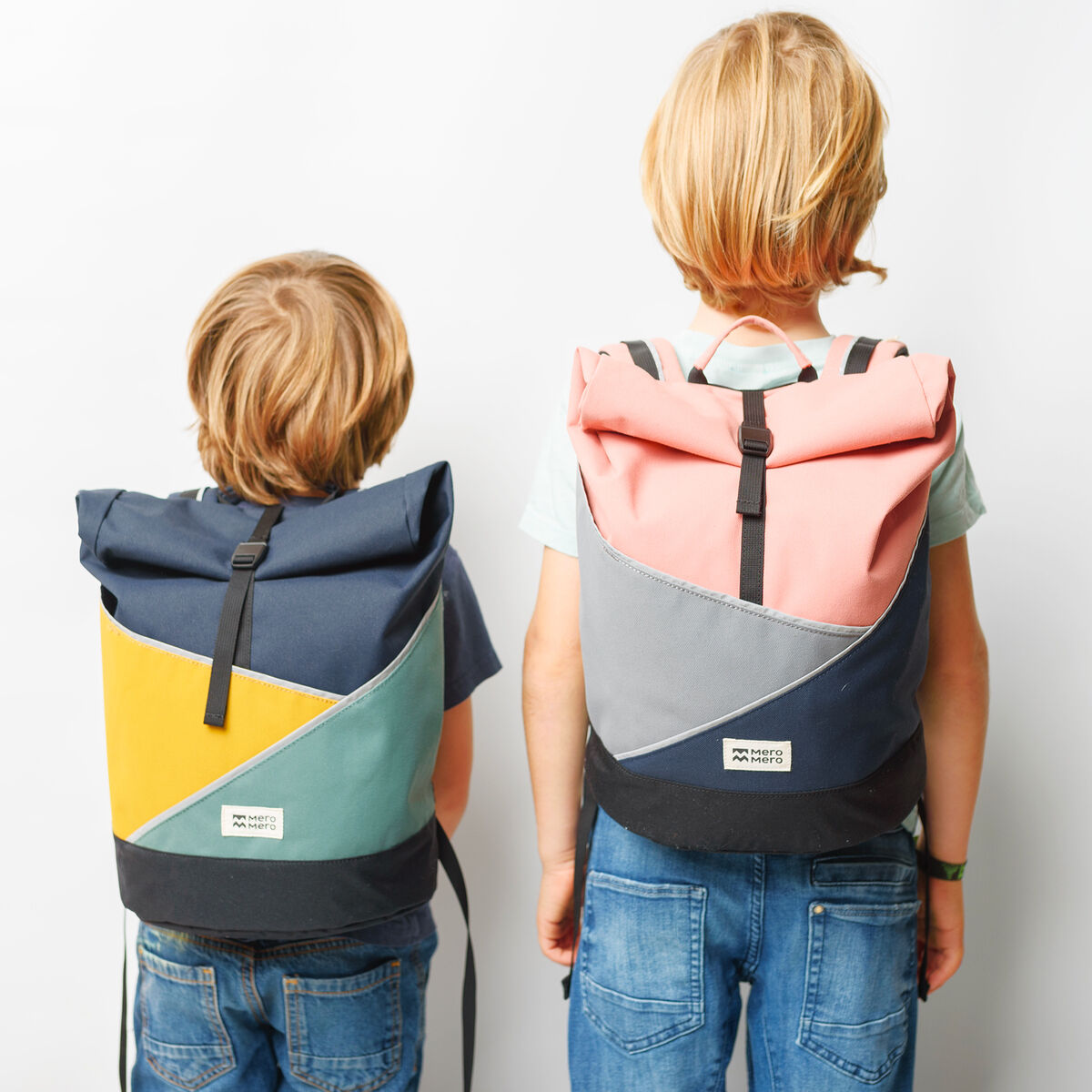 Zwei Kinder tragen bunte Rucksäcke der Marke »Popoyo« auf dem Rücken. Die Rucksäcke sind in verschiedenen Farben wie Blau, Rosa, Gelb, Weiß, Grau und Schwarz gestaltet. Eine schlichte weiße Wand bildet den Hintergrund.