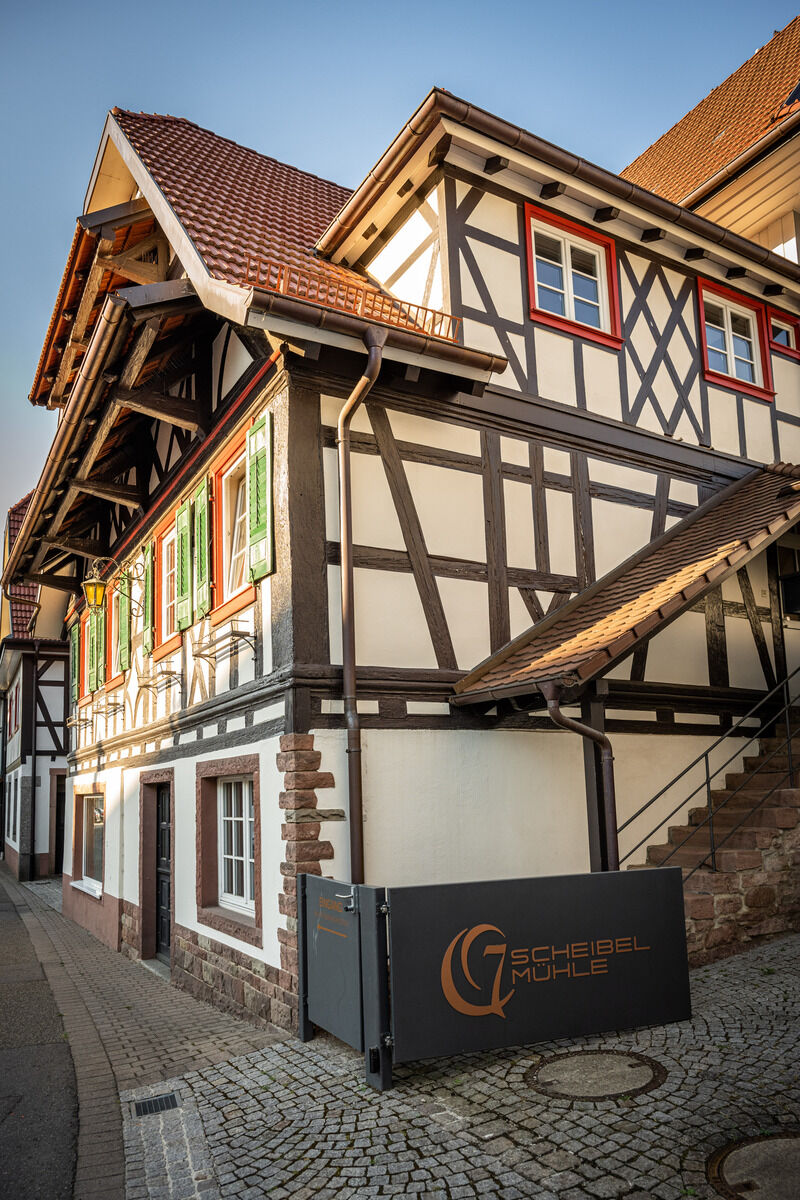 Senkrechte Aufnahme eines historischen Fachwerkhauses in einer engen gepflasterten Gasse. Weiße Gefache, dunkelbraune Holzbalken und rot gerahmte Fenster prägen die mehrstöckige Fassade mit ziegelrotem Dach. Vorn steht ein schwarzes Schild mit goldenem Logo und der Aufschrift „Scheibel Mühle“.