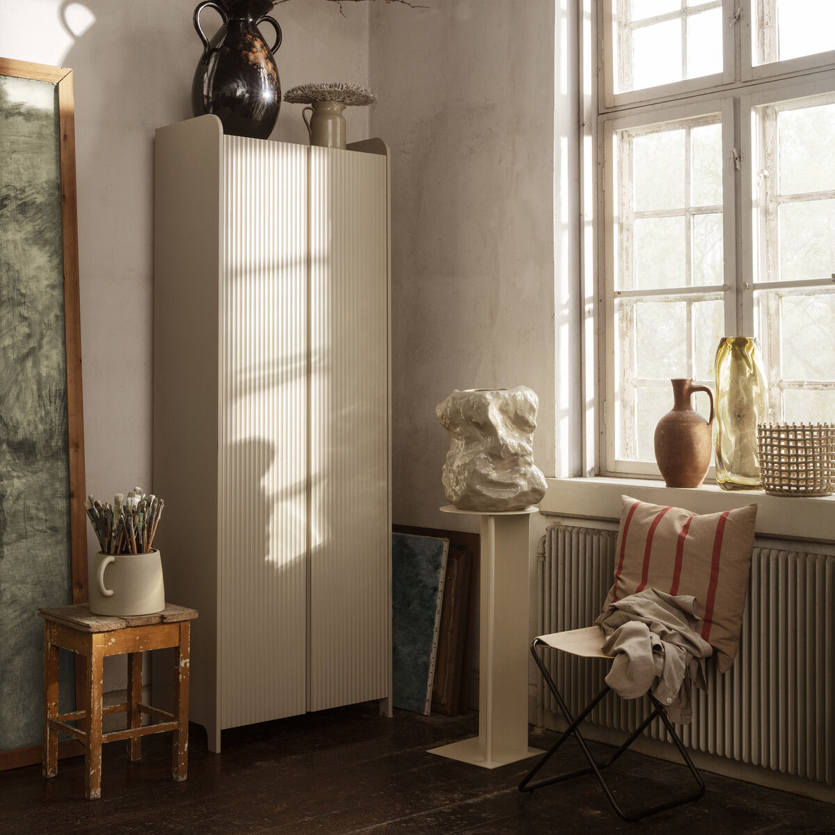 Ein stilvoll eingerichtetes Wohnzimmer mit einem Kissen 'Grand' aus Baumwolle und Leinen auf einem Stuhl. Das Zimmer hat eine beige Farbpalette mit weißen und braunen Akzenten. Große Fenster lassen viel Licht herein und es gibt verschiedene Dekorationselemente wie Vasen und Blumenarrangements.