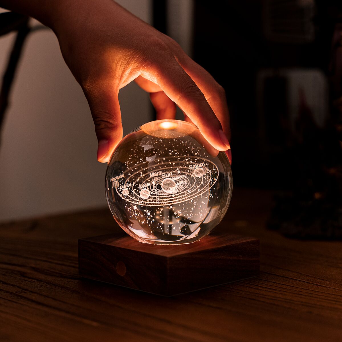 Eine kunstvoll gravierte Kristallkugel mit Sonnensystem-Motiv ruht auf einem eleganten, dunklen Walnussholzsockel. Eine Hand berührt sanft die leuchtende Kugel, die auf einem Holz-Tisch steht. Die warme Lichtstimmung schafft eine gemütliche Atmosphäre.