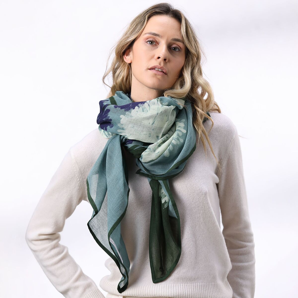 Eine Frau trägt einen großen, weichen Wollschal in Eisblau mit grünen und dunklen Farbakzenten über einem hellen Pullover. Der Schal ist locker um den Hals drapiert und passt zu einem modernen, winterlichen Look. Das Produkt steht im Vordergrund und vermittelt ein Gefühl von Wärme und Gemütlichkeit.