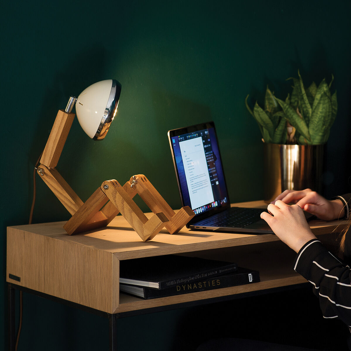 Eine Person sitzt an einem Schreibtisch mit einer Leuchte »Mr. Wattson« neben einem Laptop und einer Pflanze, im Hintergrund eine grüne Wand.