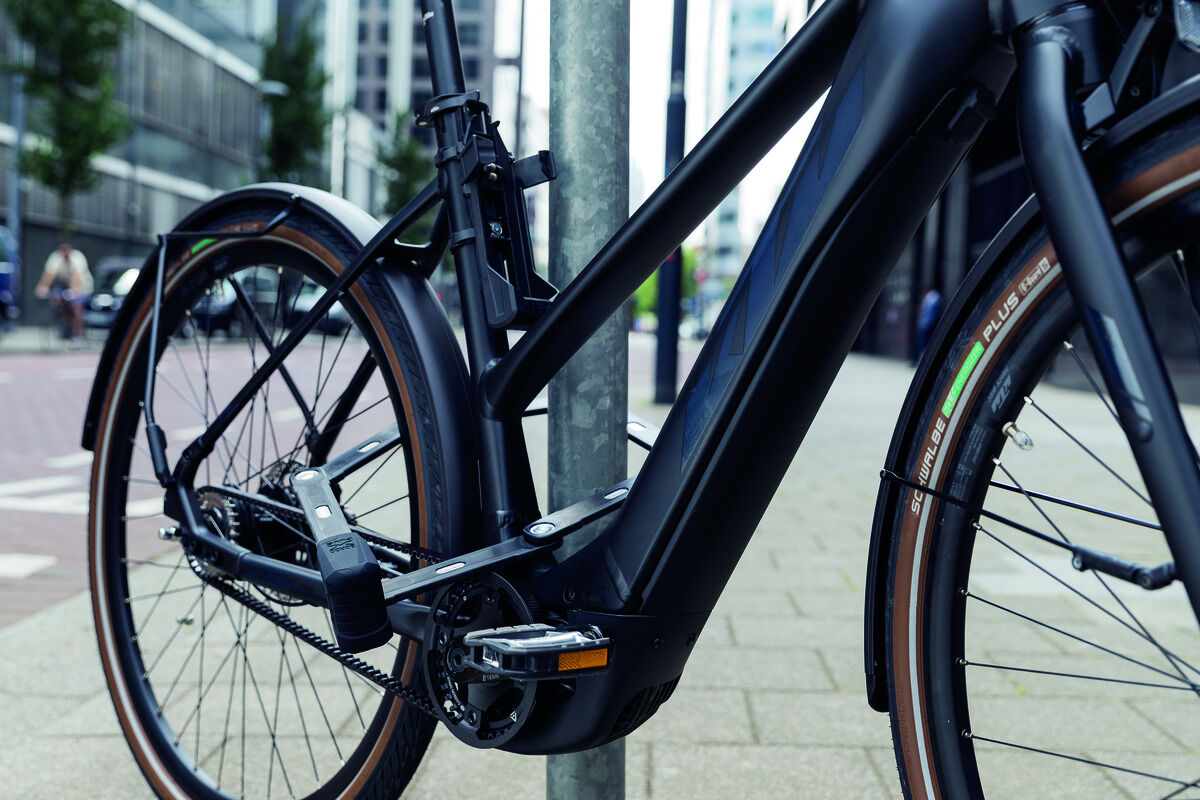 Ein modernes schwarzes Fahrrad ist mit einem Fahrrad-Faltschloss »BORDO GRANIT XPlus 6500« an einem Metallpfosten gesichert. Das Schloss zeigt eine robuste und kompakte Bauweise. Im Hintergrund sind Gebäude und ein gepflasterter Gehweg zu sehen.