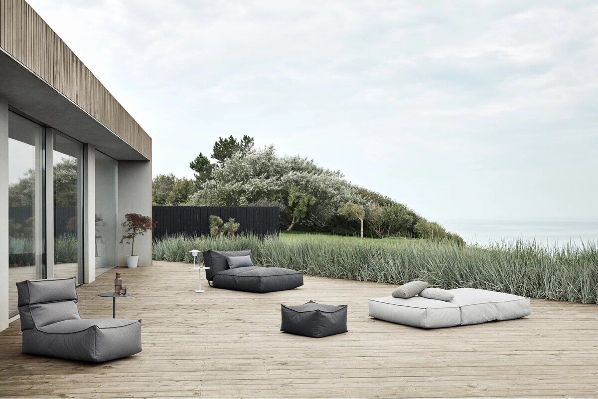 Eine moderne Outdoor-Lounge-Gruppe mit dem Produkt 'Outdoor Lounger »STAY«' auf einer Holzterrasse vor einem minimalistischen Gebäude. Die Loungemöbel sind in verschiedenen Grautönen gehalten und umgeben von grüner Vegetation und Strandlandschaft.