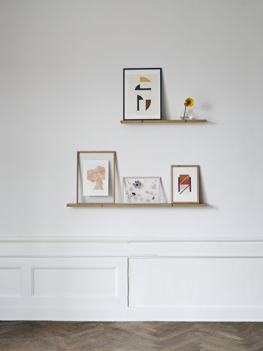 Ein minimalistisches Wandregal aus Eichenholz, das zwei dünne Regalbretter umfasst. Darauf befinden sich mehrere Bilderrahmen mit abstrakter Kunst und eine kleine Sonnenblume in einer Vase. Die Wand ist weiß gestrichen und der Boden besteht aus braunem Parkett.