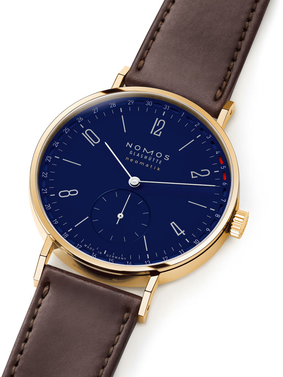 Die abgebildete Armbanduhr ist die ZEITmagazin-Sonderedition Tangente Gold neomatik 38 Update von NOMOS. Gehäuse aus 18 Karat Gold, tiefblaues Zifferblatt mit feiner roter Datumsanzeige, braunes Lederarmband. Ästhetisches, elegantes Design.