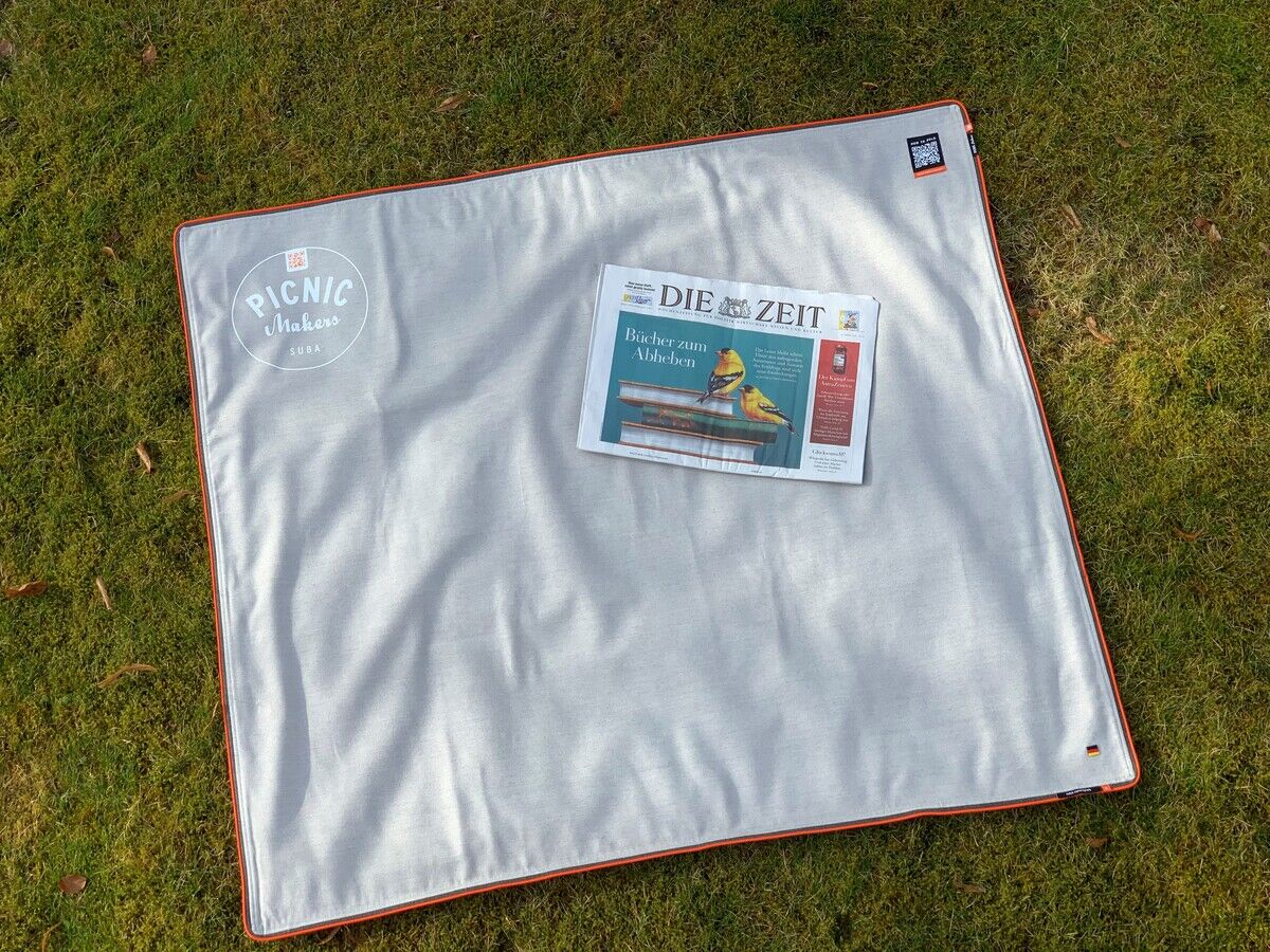 Eine Picknickdecke namens »Squarepark 11« liegt auf einer Wiese. Die Decke ist grau mit orangen Nähten und einem weißen runden Logo in der Ecke. Auf der Decke liegt eine Ausgabe der Zeitung 'DIE ZEIT'.