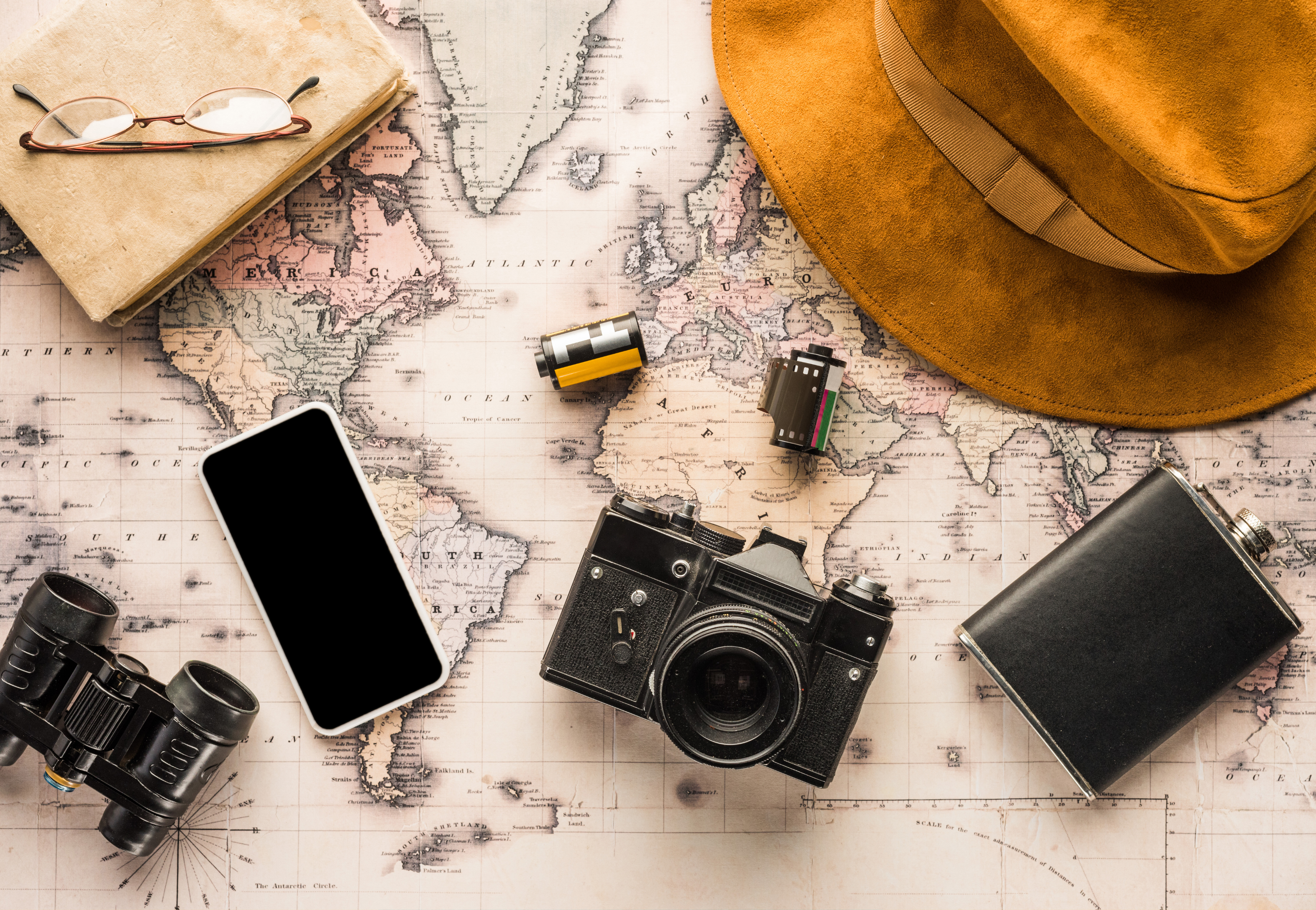 Link zur Kategorie Bücher. Eine Weltkarte als Untergrund, darauf verteilt eine analoge Kamera, ein Smartphone, ein Fernglas, ein Notizbuch mit Brille, eine Filmrolle, ein schwarzer Flachmann und ein brauner Hut.