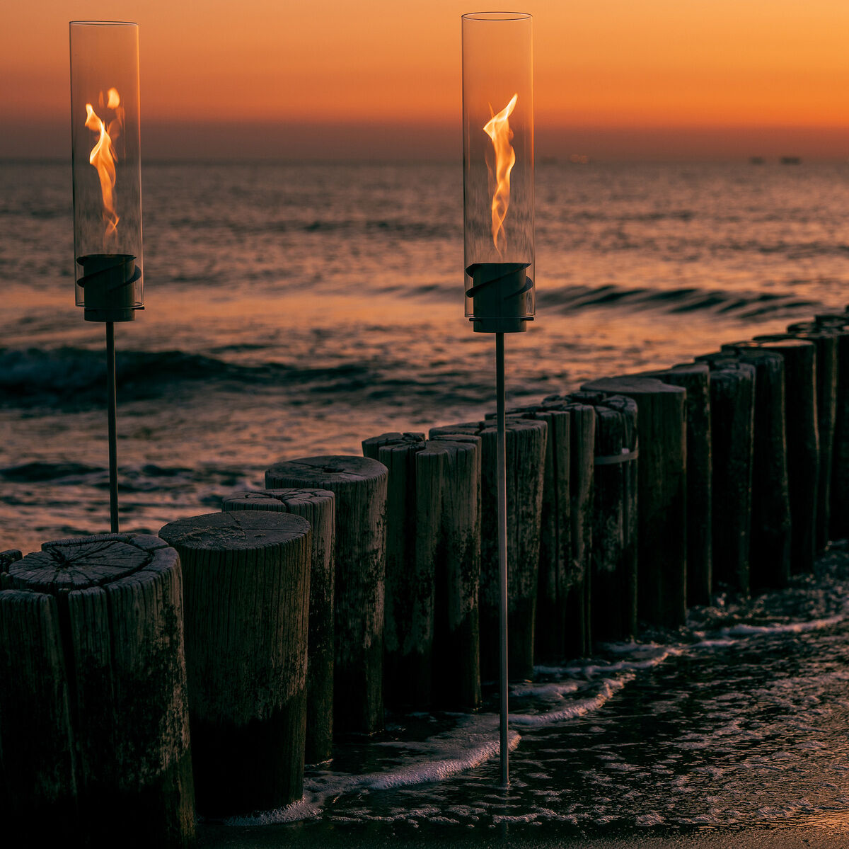 Zwei SPIN Erdspieß Fackeln leuchten am Strand bei Sonnenuntergang. Die Flammen reflektieren im Wasser und erzeugen eine romantische Atmosphäre.