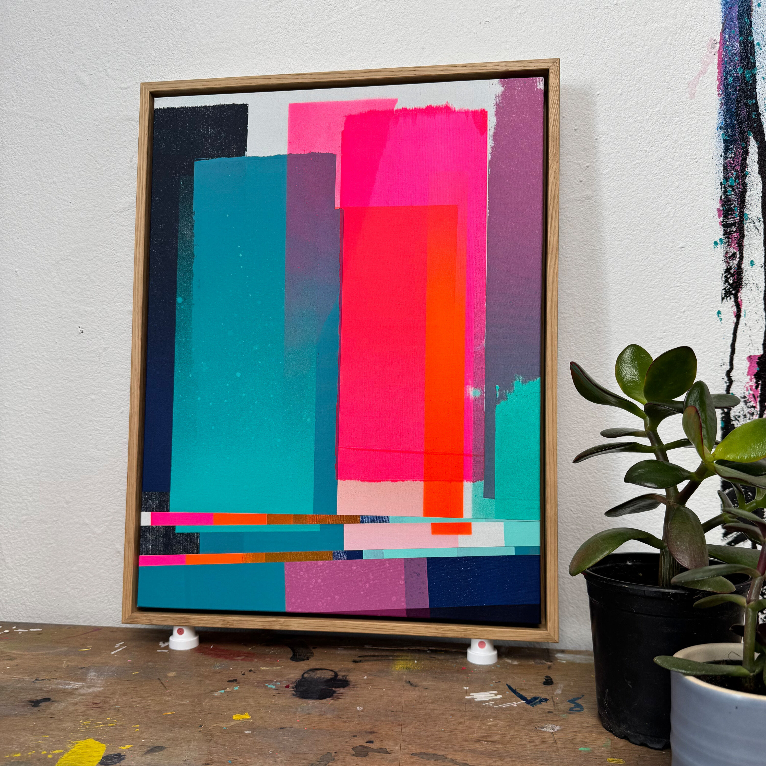 Ein Siebdruck-Kunstwerk in abstraktem Stil mit kräftigen, übereinanderliegenden Farbfeldern in Türkis, Pink, Magenta, Weiß, Dunkelblau, Orange und Violett. Das Bild ist in einem schlichten Holzrahmen gefasst und steht auf einer Werkbank vor einer weißen Wand. Rechts im Vordergrund ist eine grüne Zimmerpflanze erkennbar.