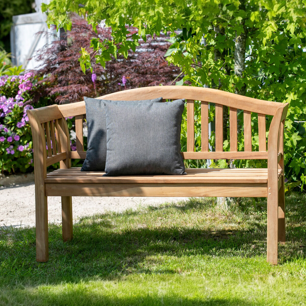 Teak-Friesenbank »Wave« von bene living im Garten: Die unbehandelte, hellbraune Holzbank hat sanft geschwungene Rücken- und Armlehnen sowie senkrechte Sprossen. Ein großes dunkelgraues Kissen liegt mittig auf der Sitzfläche. Sie steht auf grünem Rasen vor dichtem Laub, violetten Blüten und einem rötlichen Strauch.