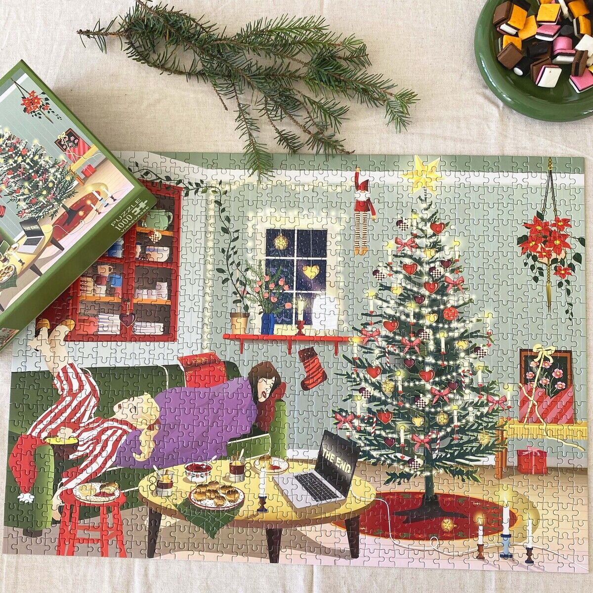 Vollständig zusammengesetztes 1000-Teile Puzzle „Faule Tage“ von VISSEVASSE zeigt ein gemütliches Wohnzimmer: Zwei Kinder liegen im Schlafanzug auf einem Sofa, Weihnachtsbaum, Geschenke, Kerzen, Laptop und Snacks sorgen für festliche Stimmung. Tannenzweig und bunte Schale daneben.