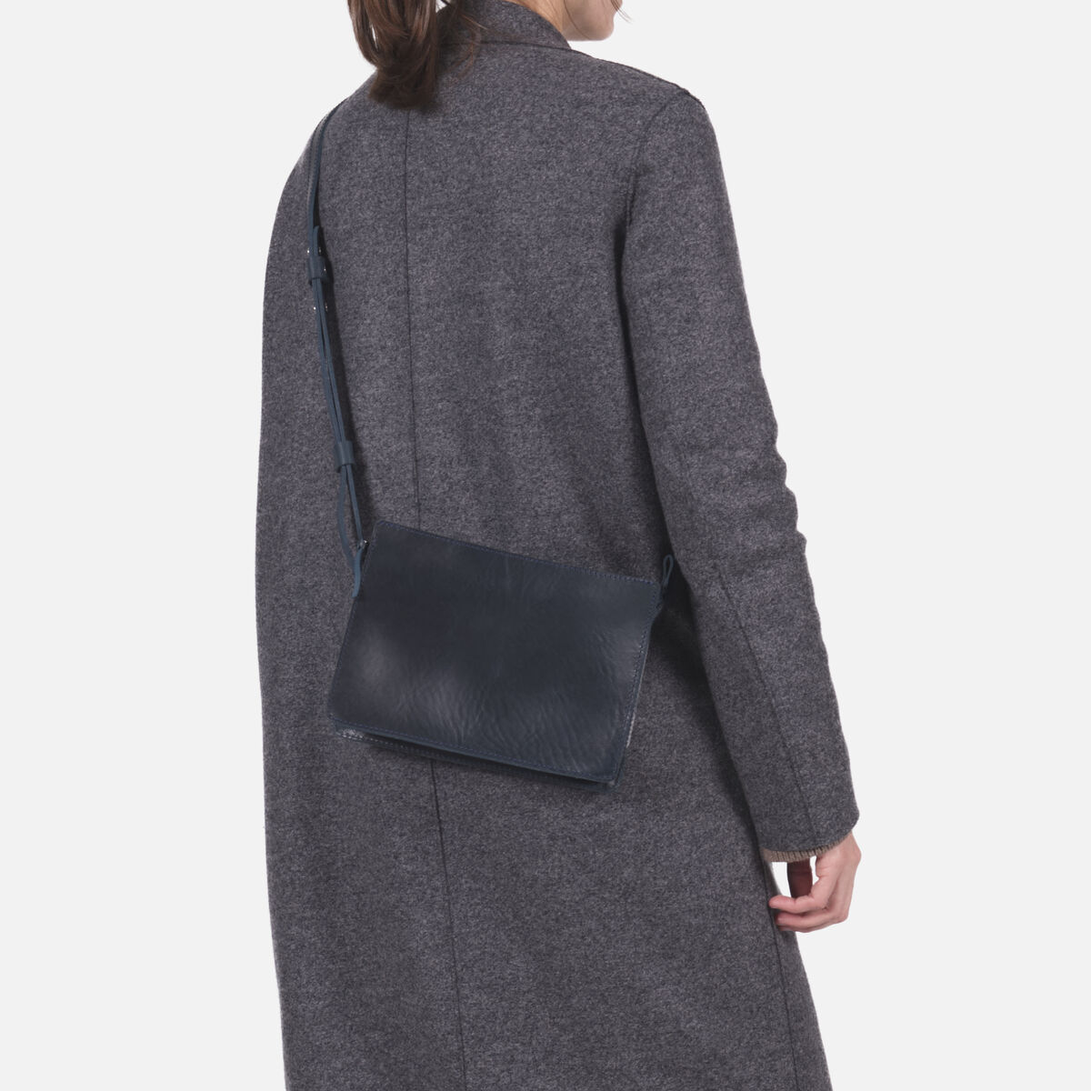 Frau mit dunkelblauer Kernleder-Umhängetasche, getragen über der Schulter, bekleidet mit grauem Mantel und hellen Hosen vor hellem Hintergrund. Die Tasche ist schlicht, rechteckig und passt zu modernen, minimalistischen Outfits.