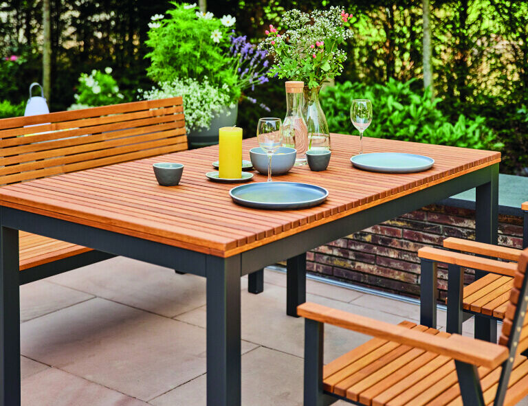 Holztisch »Bozen« in Größe 160x90cm auf einer Terrasse. Der Tisch ist aus braunem Holz mit schwarzen Metallbeinen. Auf dem Tisch befinden sich vier Teller, ein gelbes Kerzenlicht, Gläser und eine Vase mit Blumen. Im Hintergrund sind Pflanzen und eine grüne Hecke zu sehen.