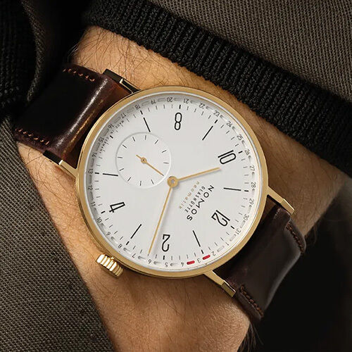 Exklusive Armbanduhr NOMOS Tangente Gold neomatik 38 Update DIE ZEIT mit goldenem Gehäuse, weißem Zifferblatt, schwarzer Typografie und roten Akzentpunkten am Ringdatum. Dunkelbraunes Lederarmband, getragen am Handgelenk, fein und modern inszeniert.