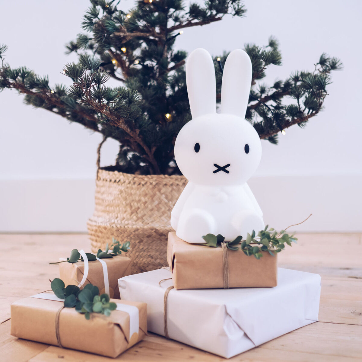 Ein Nachtlicht in Form von Miffy, einem weißen Kaninchen, steht auf einem Holzboden vor einem kleinen Weihnachtsbaum. Umschlossen von braunem und weißem Papier verpackte Geschenke, teilweise dekoriert mit grünen Zweigen, liegen um das Nachtlicht herum.