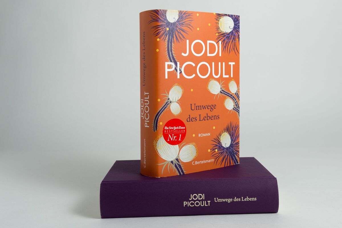 Das Bild zeigt das Buch 'Umwege des Lebens' von Jodi Picoult in gebundener Ausgabe. Das Cover ist orange mit weißen und blauen Blumenmotiven und zeigt den Namen der Autorin in großen weißen Buchstaben. Ein rotes Siegel auf dem Cover deutet darauf hin, dass es ein Bestseller ist. Das Buch liegt auf einem lila Buchrücken, der ebenfalls den Titel und den Namen der Autorin in weißen Buchstaben trägt.