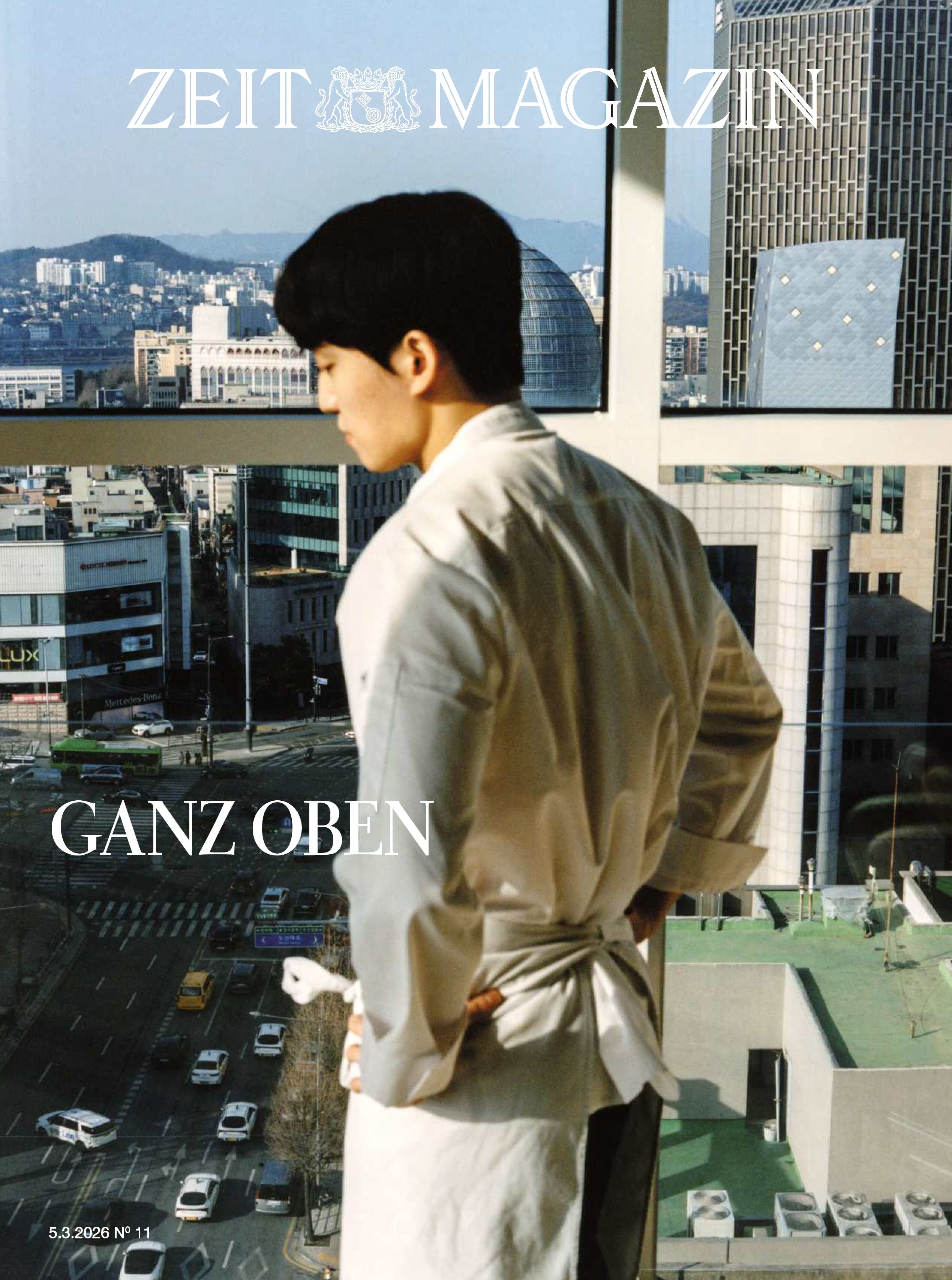 Titelcover des ZEIT MAGAZIN Nr. 11 von 2026. Ein junger Mann in weißer Kochjacke steht an einem großen Fenster mit Blick auf eine Großstadt mit Hochhäusern, Straßen, Autos und Bergen im Hintergrund. Aufschrift: „GANZ OBEN“.