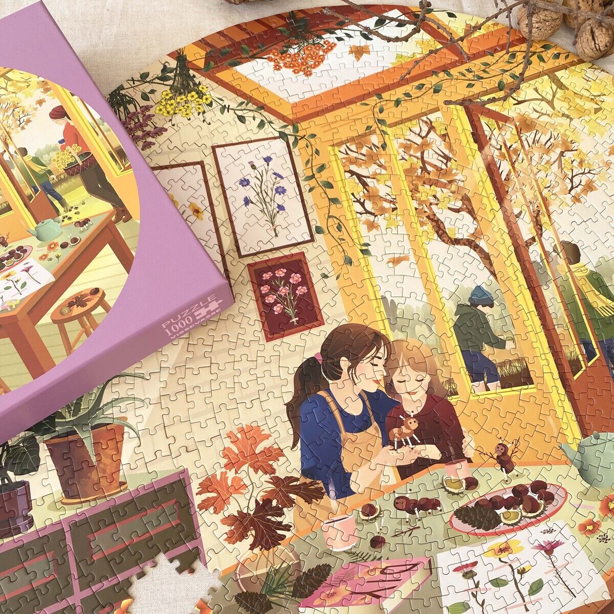 Zu sehen ist das runde 1000-Teile Puzzle »Chestnut Season« von VISSEVASSE. Das Motiv zeigt eine herbstliche Indoor-Szene mit zwei zeichnenden Personen am Tisch, umgeben von Pflanzen und Fenstern mit gelb-orangen Farbtönen. Links liegt der lilafarbene Karton mit Motivbild.