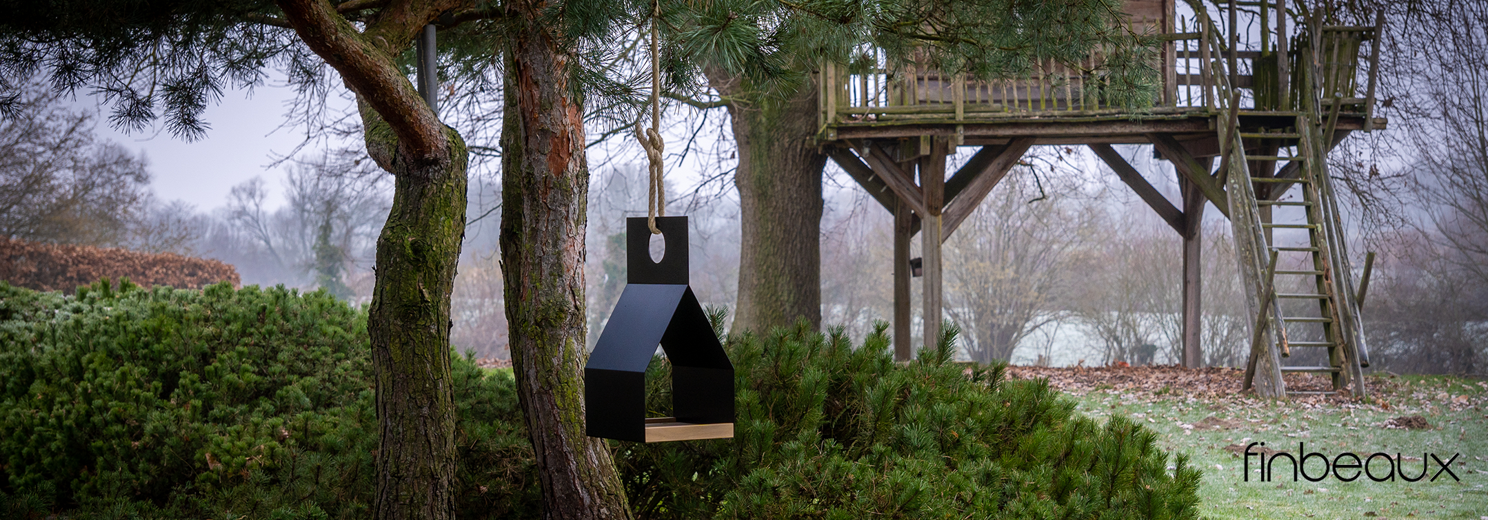 Schwarze, hängende Skulptur aus geometrischen Formen vor Bäumen und einem Holzbaumhaus mit Leiter