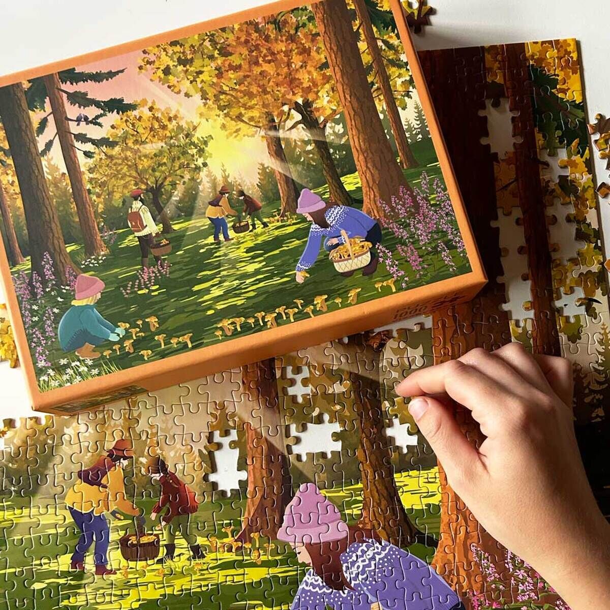 Das Bild zeigt ein fast fertig zusammengesetztes 1000-Teile Puzzle mit einer herbstlichen Waldszene: Mehrere Personen sammeln in einem sonnenbeschienenen Laubwald Pilze. Im Vordergrund ist eine Hand zu sehen, die an dem Puzzle arbeitet. Das Motiv besticht durch warme Grün- und Gelbtöne sowie violette Akzente. Die Puzzleschachtel mit dem gleichen Motiv liegt daneben.