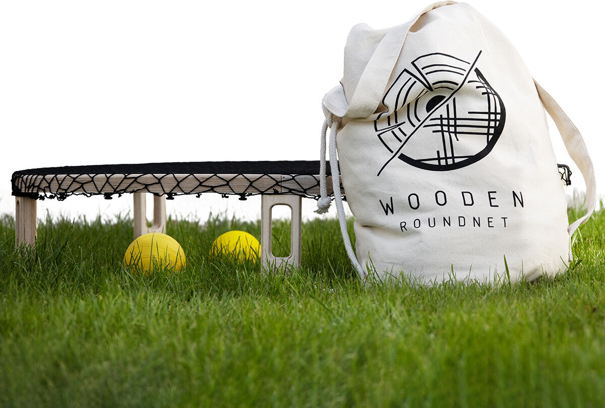 Ein Wooden Roundnet Set bestehend aus einem runden Netz auf Holzfüßen, mehreren Bällen und einer weißen Tragetasche mit Logo, auf grünem Gras platziert.