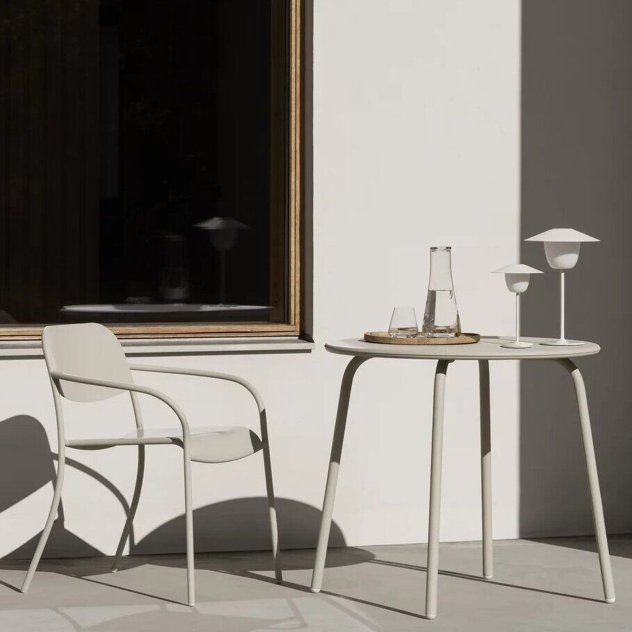 Eine moderne, mobile LED Tischleuchte mit dem Namen 'ANI' steht auf einem weißen, minimalistischen Tisch neben einem passenden Stuhl. Die Szene ist draußen, möglicherweise auf einer Terrasse oder Veranda, mit einer ruhigen und friedlichen Atmosphäre. Im Hintergrund ist ein großes Fenster mit Holzrahmen sichtbar.