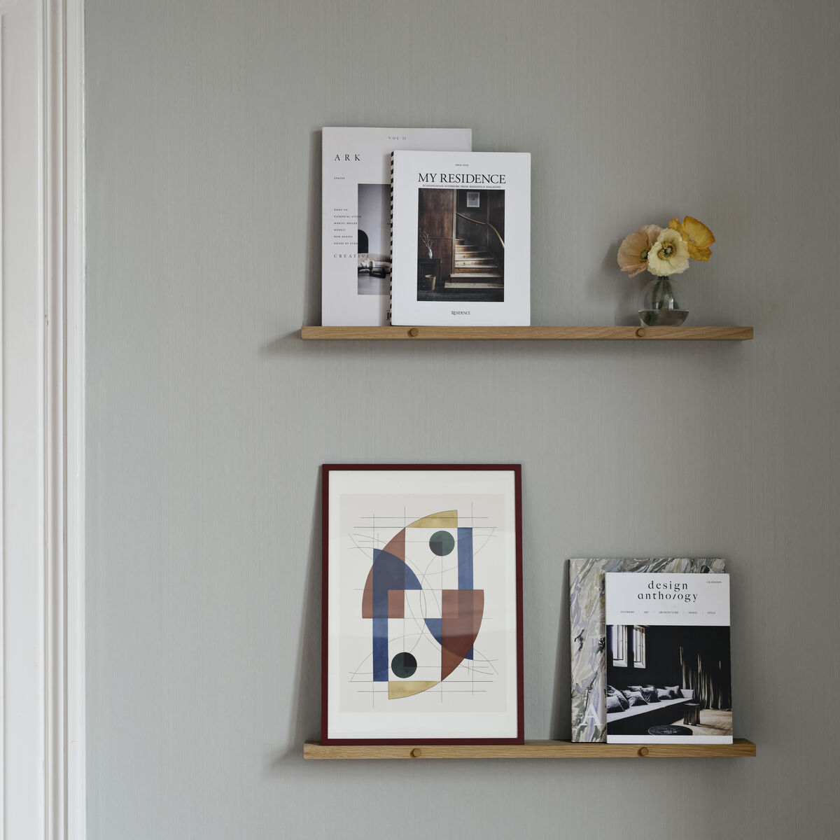 Zwei schwebende Regale aus Eichenholz an einer grauen Wand. Auf dem oberen Regal stehen Bücher und ein kleiner Blumenstrauß. Das untere Regal zeigt ein Kunstwerk und weitere Bücher.