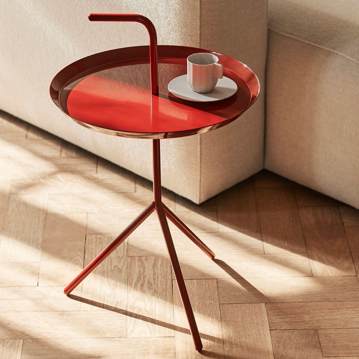 Ein moderner Beistelltisch mit runder, roter Tischplatte und einem roten Standfuß aus Eisen, der in einem dreibeinigen Design endet. Auf dem Tisch steht eine kleine weiße Tasse. Der Tisch steht auf einem Holzfußboden und neben einem beigen Sofa.