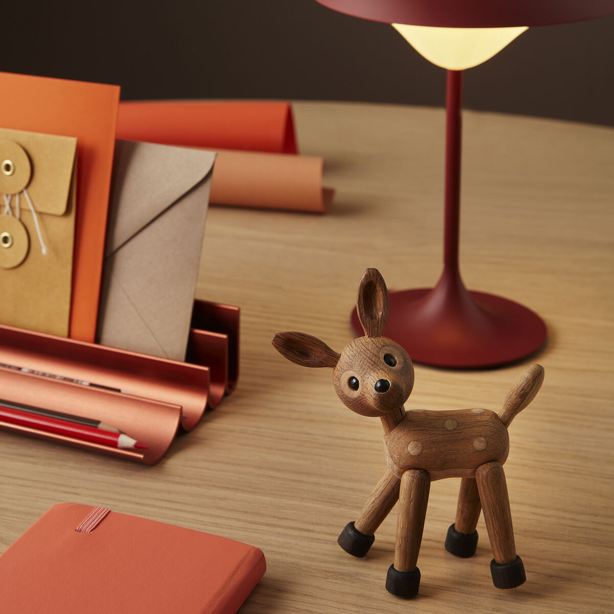 Auf einer hellen Holzoberfläche steht eine kleine, detailreich gearbeitete Holzfigur in Form eines Rehkitzes mit schwarzen Füßen und Punkten auf dem Rücken. Daneben befinden sich orangene Notizbücher, Briefumschläge, Bleistifte und eine rote Tischlampe mit rundem Schirm. Der Hintergrund ist dezent, stimmig in warmen Farbtönen gehalten und schafft eine gemütliche Atmosphäre.