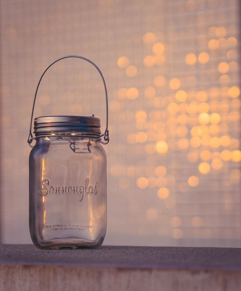 Ein Sonnenglas Classic, eine Beleuchtung in Form eines Einmachglases mit einem Solardeckel und Metallhenkel, steht auf einer Oberfläche. Im Hintergrund sind unscharfe goldene Lichtpunkte sichtbar.