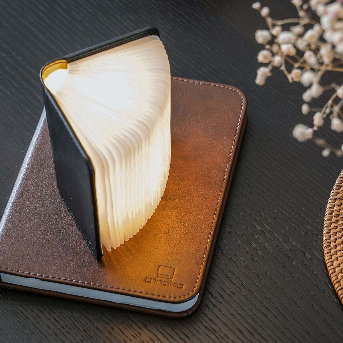 Ein Smart Booklight in Schwarz, das wie ein aufgeklapptes Buch aussieht, steht auf einer braunen Lederoberfläche. Die Seiten des Buches sind weiß und erleuchtet. Im Hintergrund sind unscharfe Elemente zu sehen, darunter ein Blumenstrauß auf der rechten Seite.